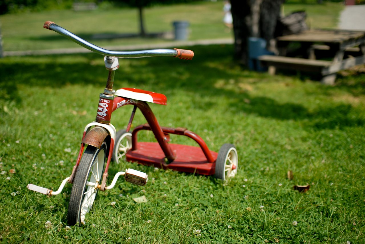 A worn red tricycle with white wheels sits on the grass in a park, slightly tilted to one side.