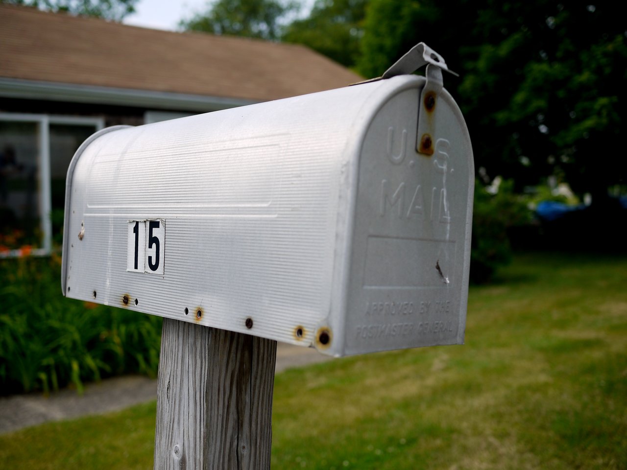 A weathered metal mailbox with the number 15, mounted on a wooden post in front of a house.