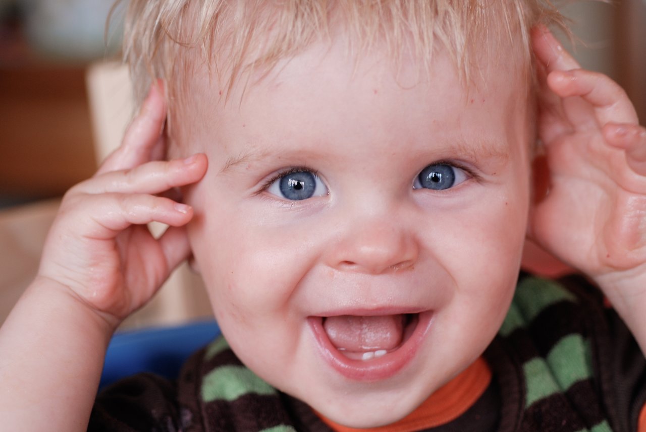 A smiling baby with blue eyes holds their hands near their head, looking directly at the camera.