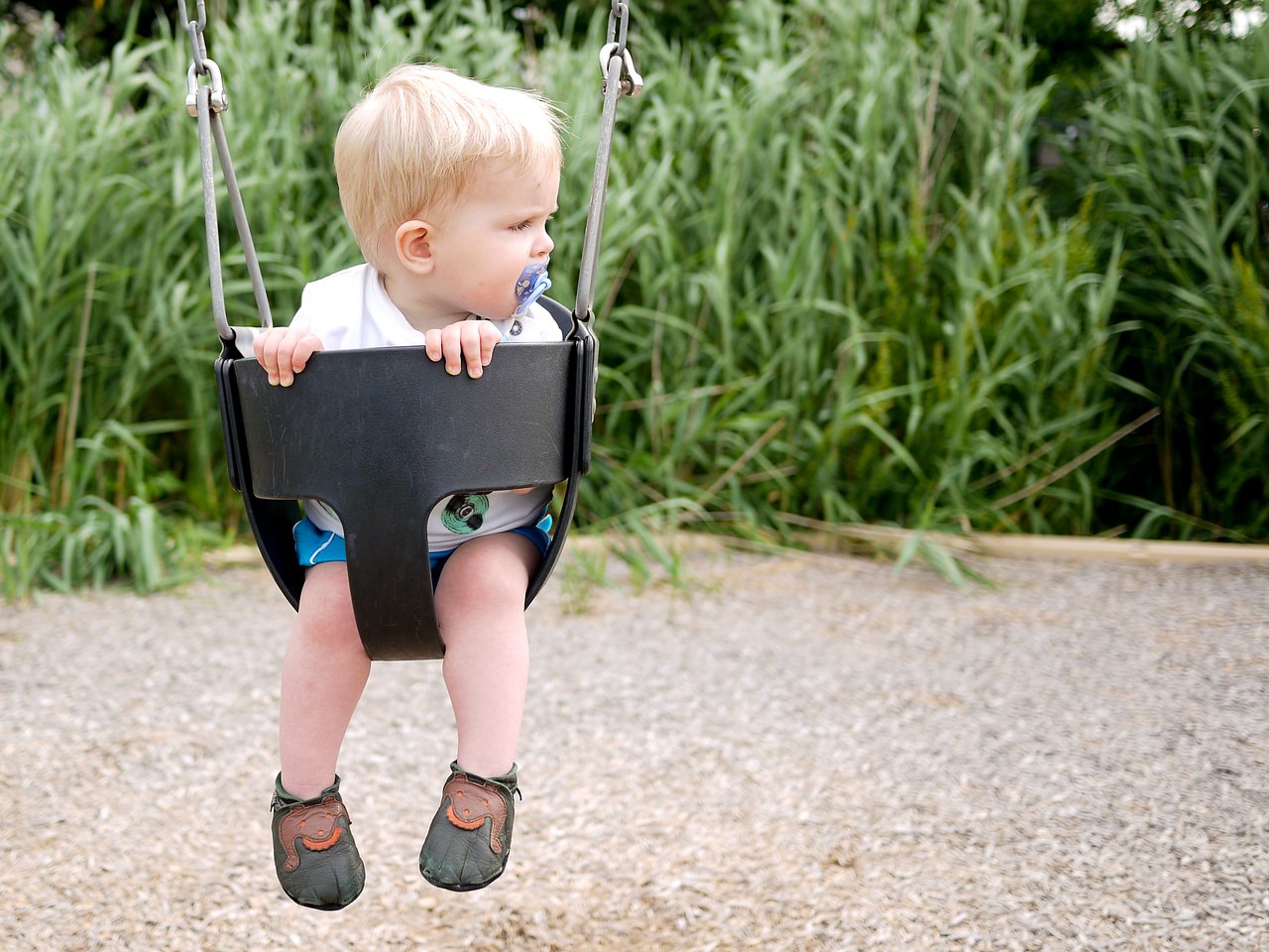 A young child sits in a swing, holding the sides and looking to the left while sucking on a pacifier.