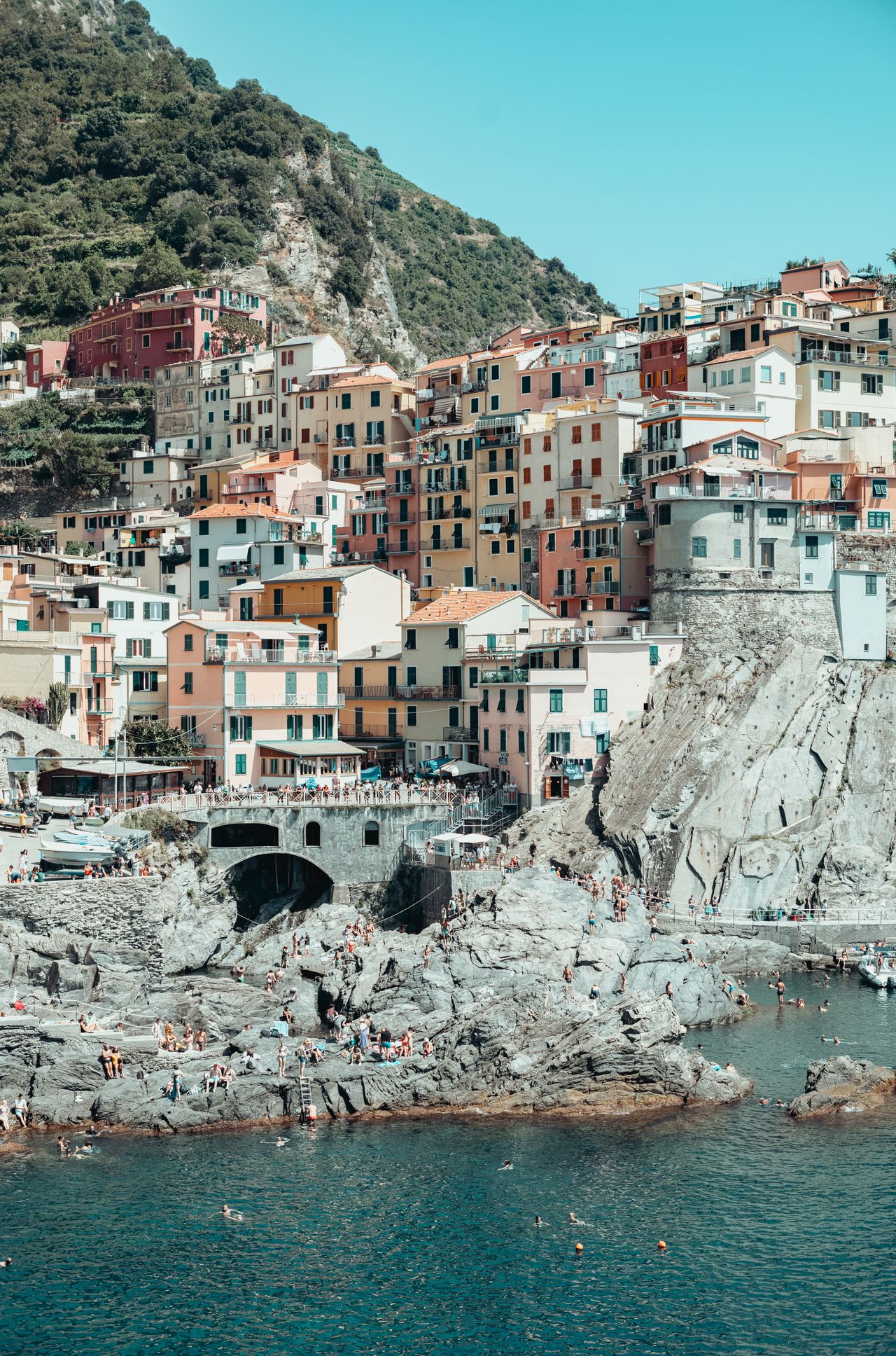 Colorful hillside buildings in Manarola, Cinque Terre, with people swimming and relaxing on rocky cliffs by the water.