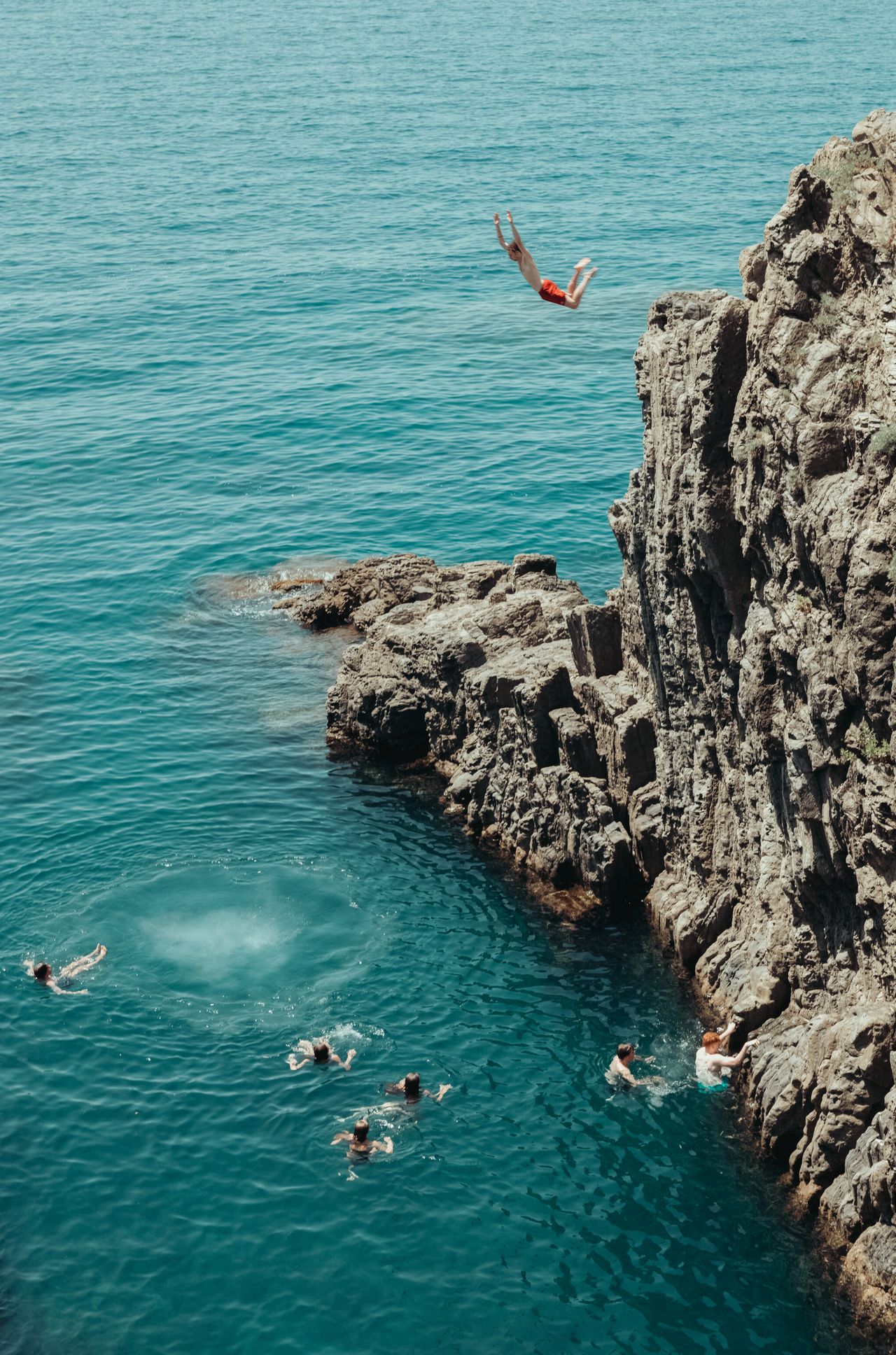A person in red shorts jumps off a rocky cliff into the sea while others swim below.