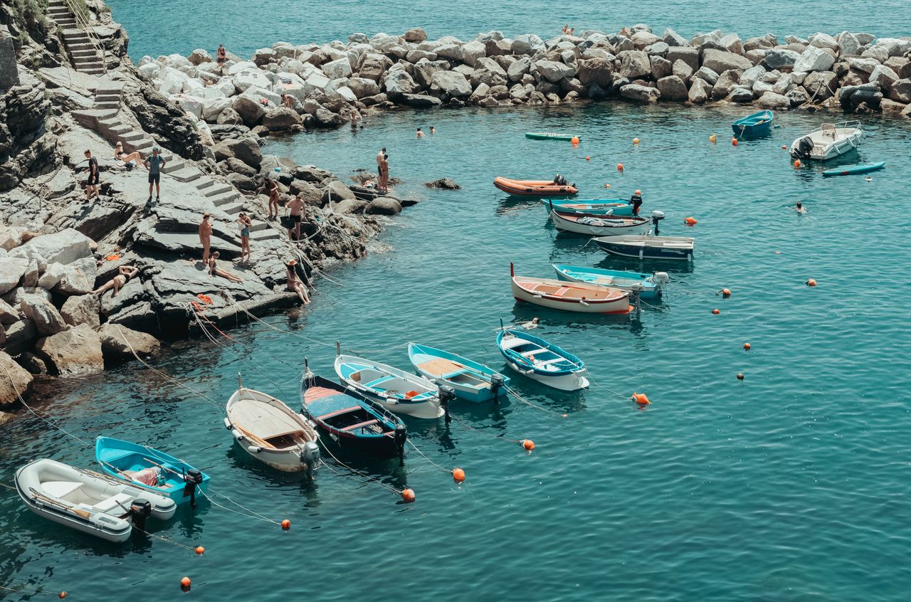 Small fishing boats floating on calm water in Riomaggiore, Cinque Terre, with people on nearby rocky steps.