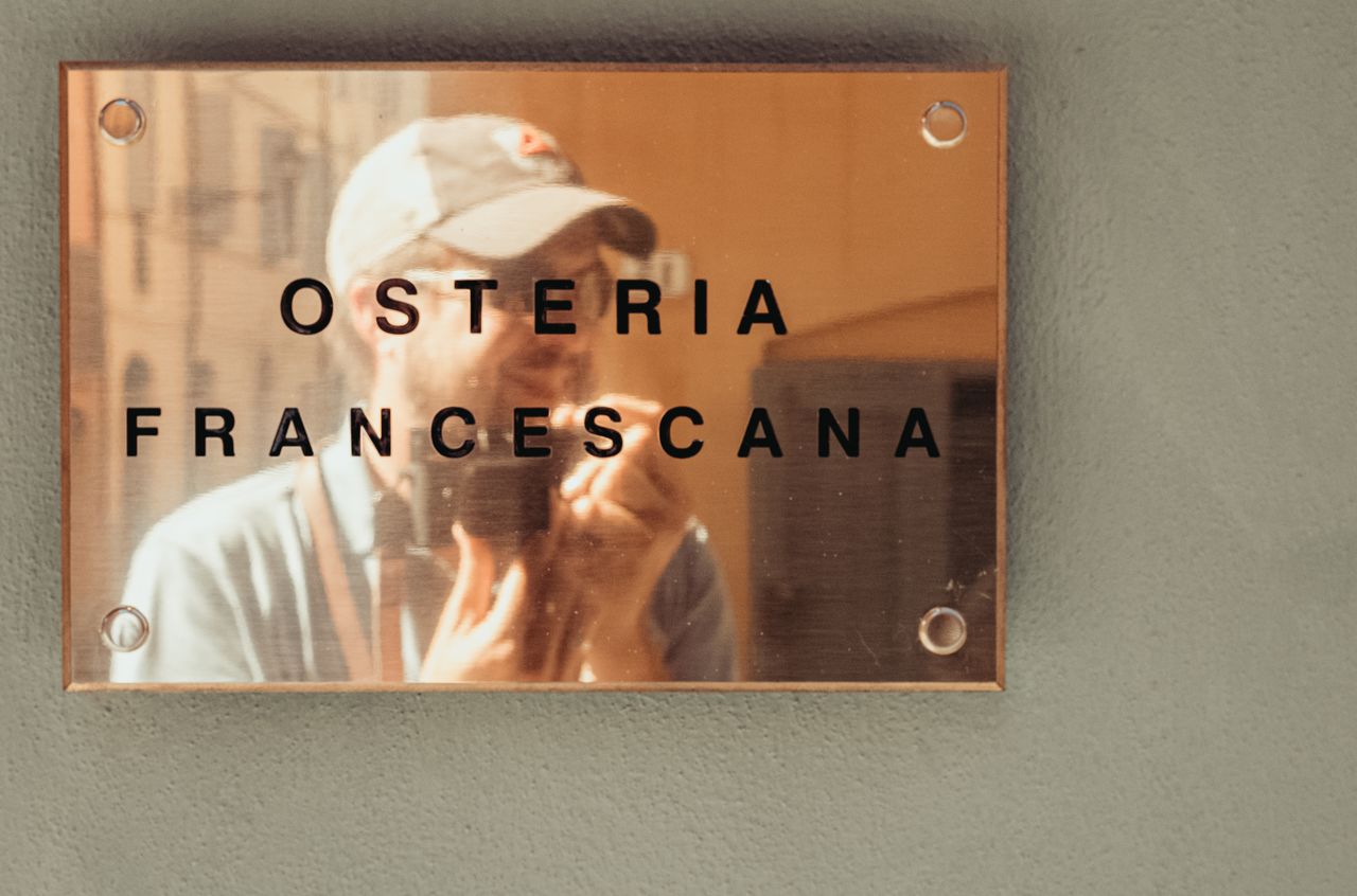 A person takes a selfie reflected in a restaurant sign that reads "Osteria Francescana.