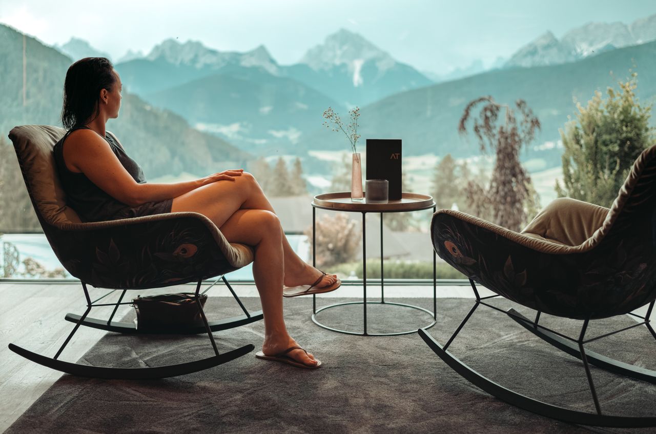 A person sits in a rocking chair, looking at mountain views through a large window at a hotel.