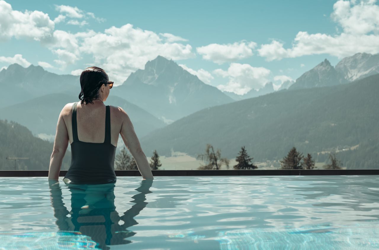 A woman in a black swimsuit stands in an outdoor infinity pool, looking at the mountains in the distance.