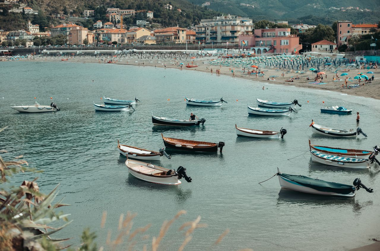 Small boats floating near the shore of Levanto beach, with people and umbrellas scattered along the sandy coastline.