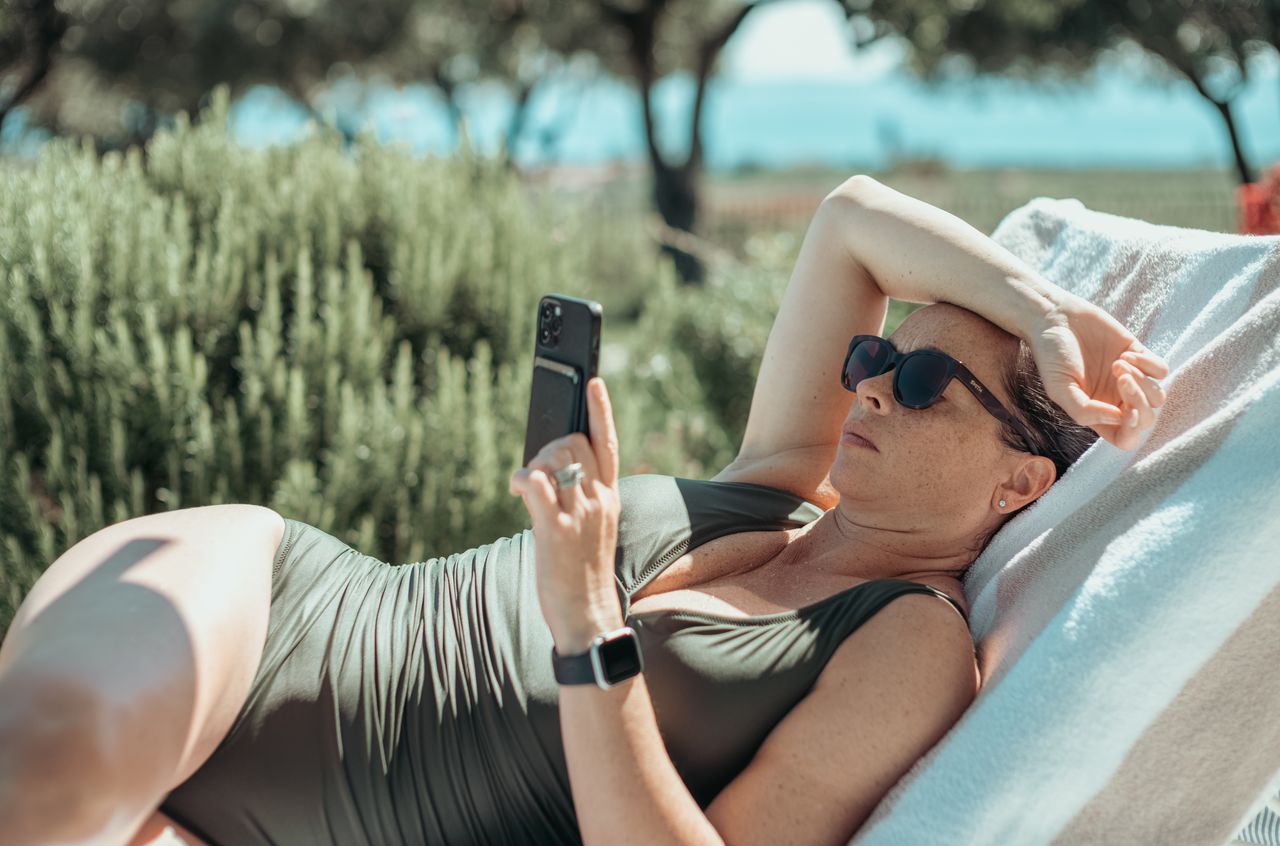 A woman in sunglasses reclines on a lounge chair by the pool, reading on her smartphone.