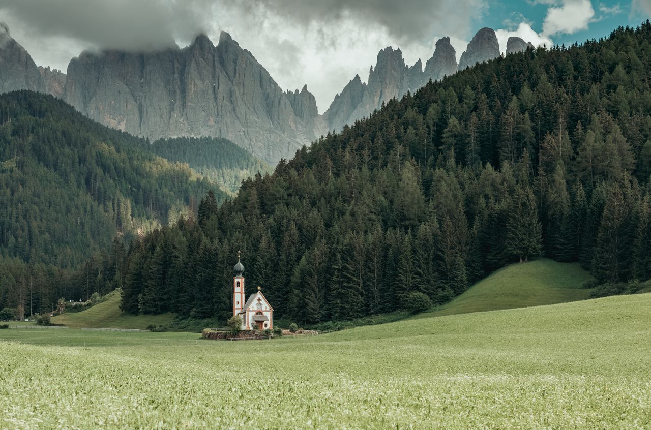 A small church with a tall steeple stands in a green valley, surrounded by trees and mountains.