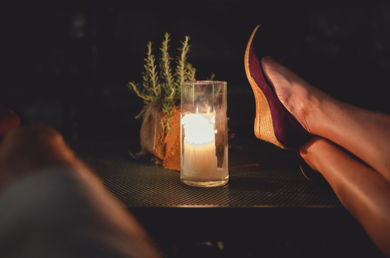 A lit candle in a glass sits on a table, with a person resting their feet nearby.
