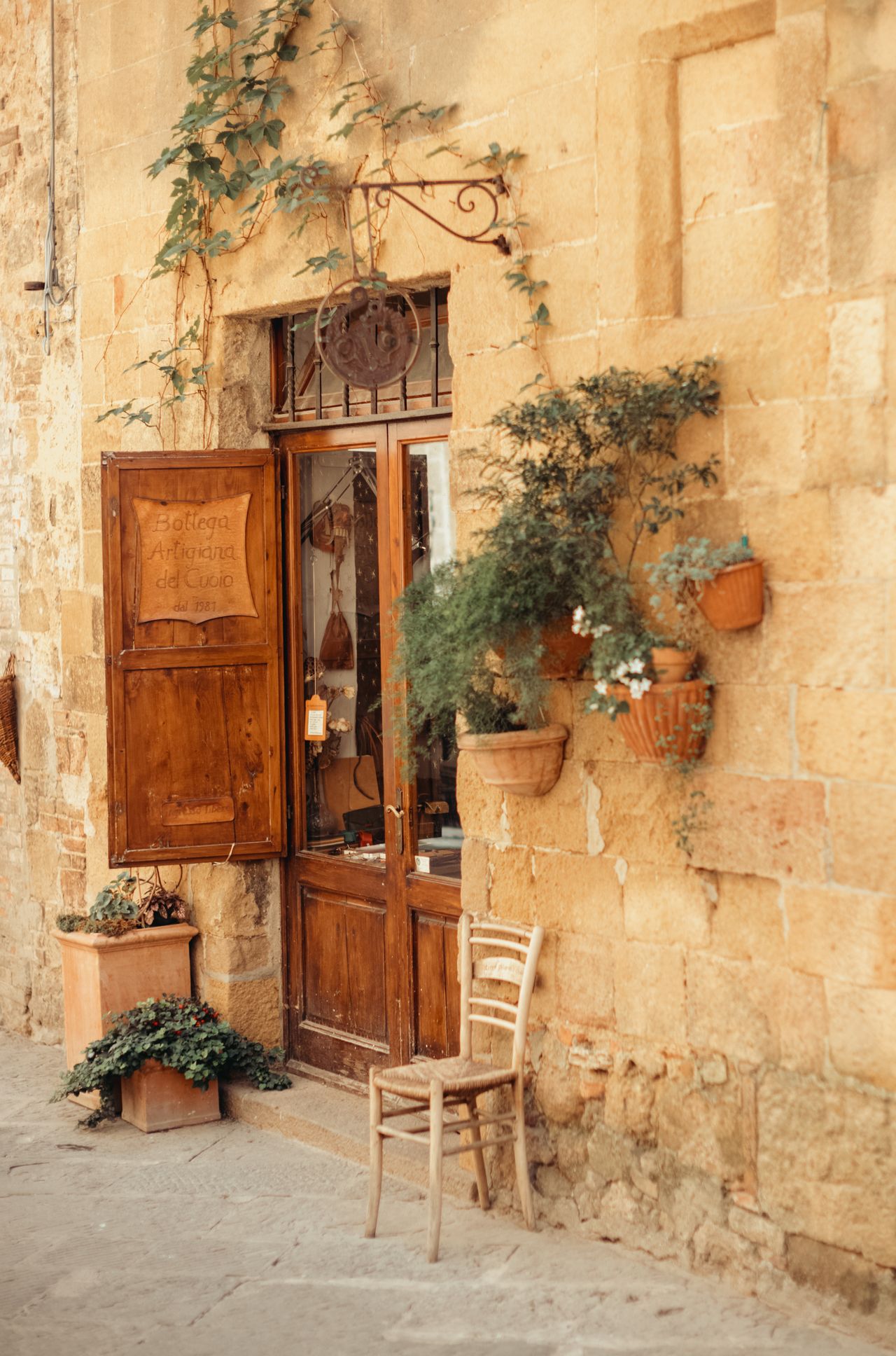 A small shop in Pienza with wooden doors, potted plants, and a chair outside on a stone street.