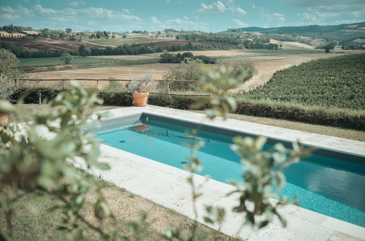 A rectangular swimming pool with clear water, surrounded by grass and plants, overlooking rolling hills and farmland.