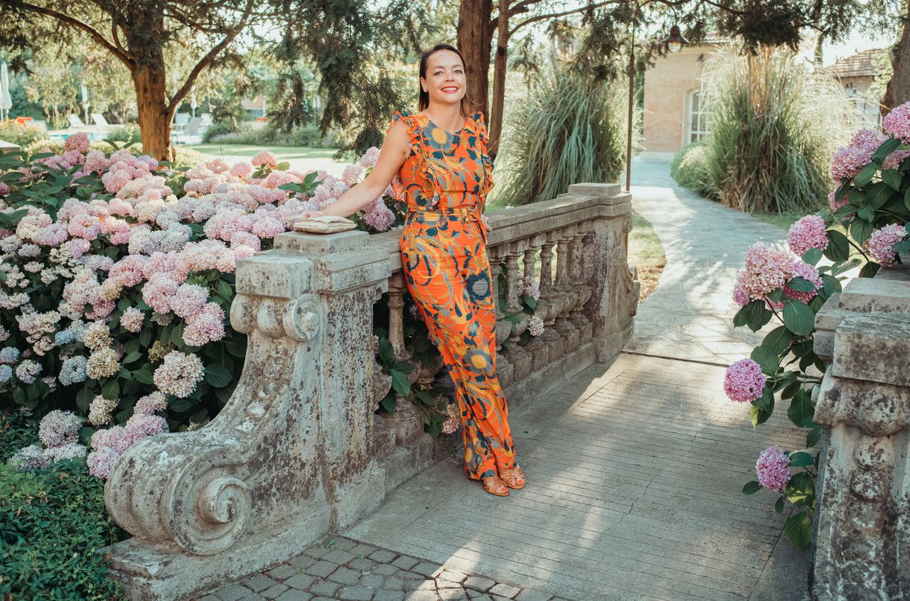 A woman in a colorful outfit leans on a stone railing, smiling, surrounded by pink flowers and greenery.