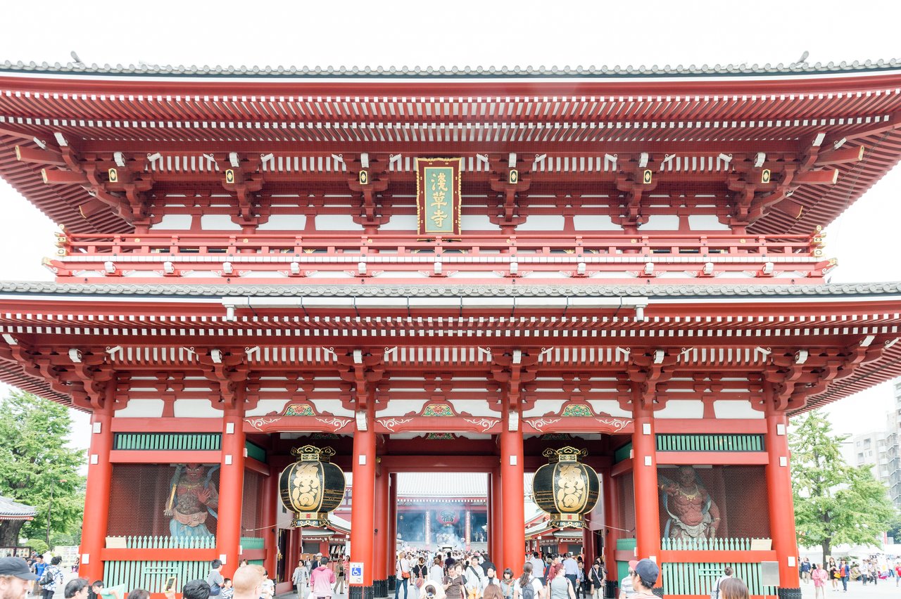 A large red temple gate with lanterns and statues, surrounded by a crowd of visitors walking through.