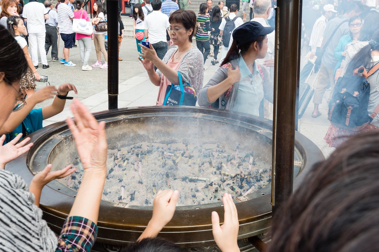 People wave incense smoke over themselves at a large incense burner in a crowded outdoor setting.