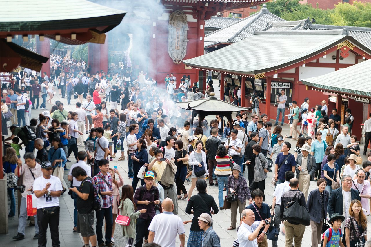 A large crowd gathers at a Japanese temple, with people walking, talking, and standing near a smoking incense burner.