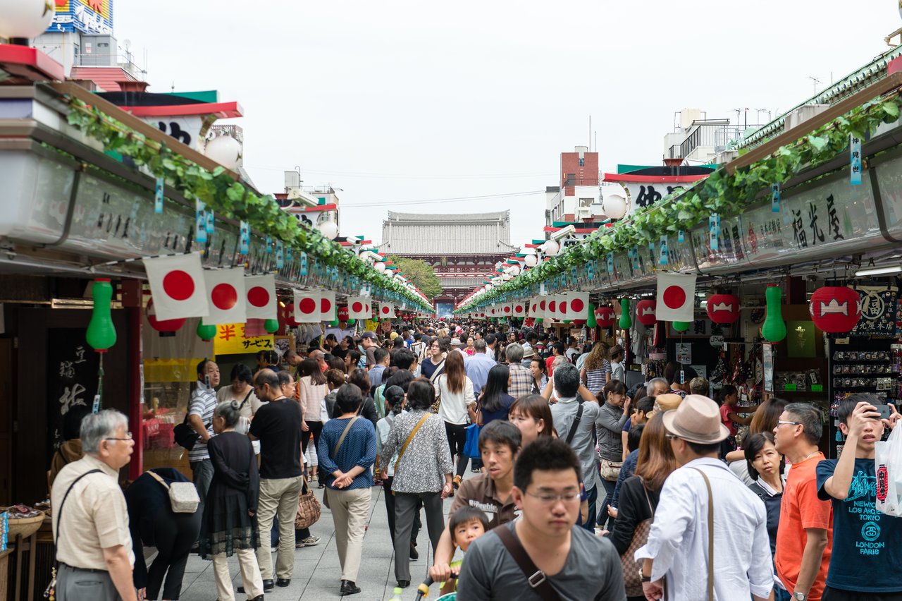 A busy outdoor market in Japan with people walking and shopping, surrounded by stalls decorated with Japanese flags.