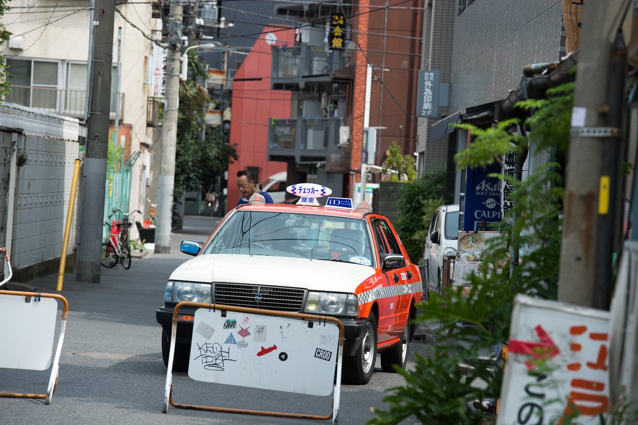 A red and white taxi is stopped behind barricades on a narrow street, with a man walking nearby.