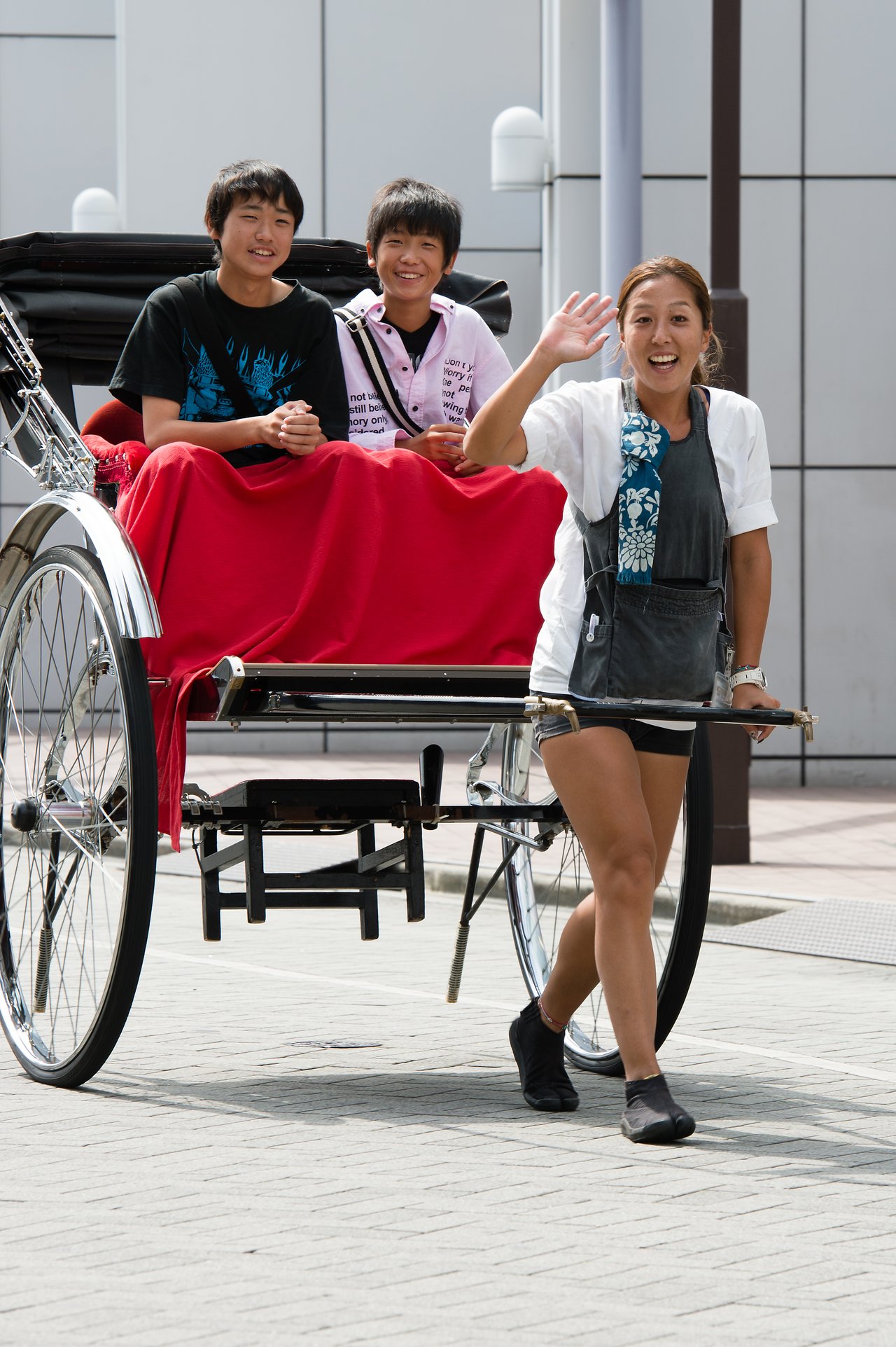 A rickshaw puller smiles and waves while pulling two passengers who are seated and smiling in the rickshaw.