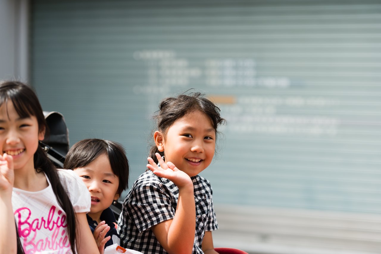 Three smiling children wave at the camera, with one girl in a checkered shirt in the foreground.