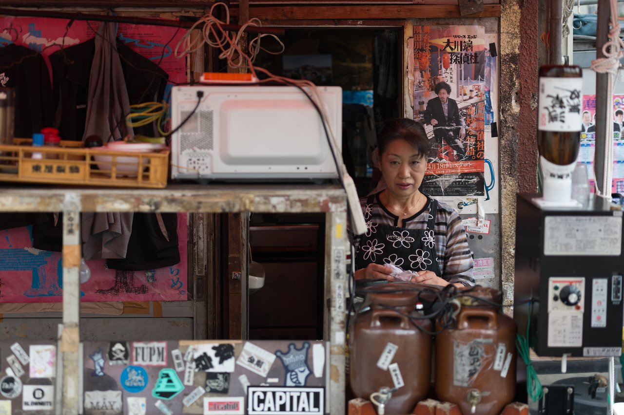 A woman in an apron stands behind a small shop counter, handling a plastic bag.