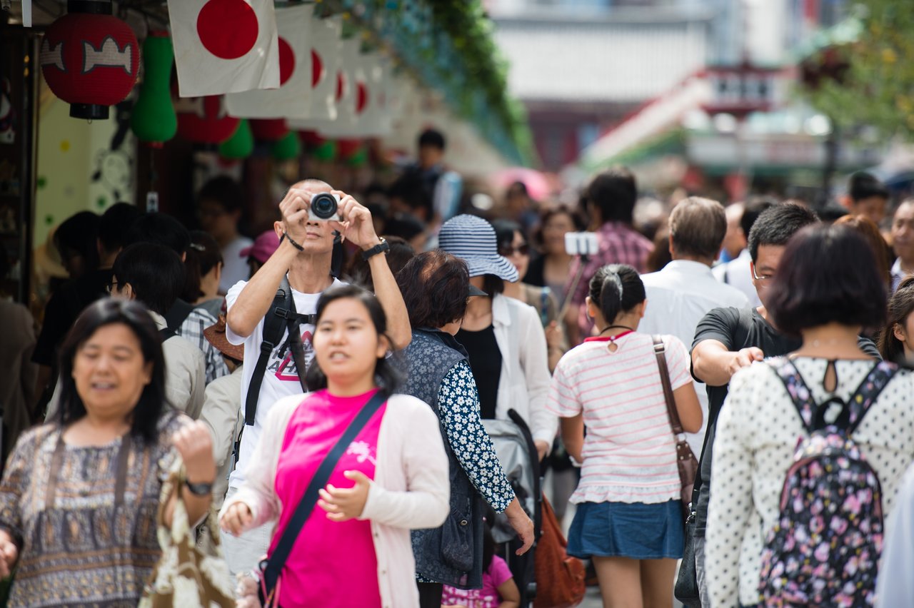 A man in a crowded street takes a photo with his camera, surrounded by people walking and shopping.