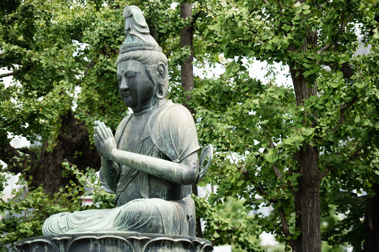 A bronze statue of a seated Buddha with hands in a prayer position, surrounded by green trees.