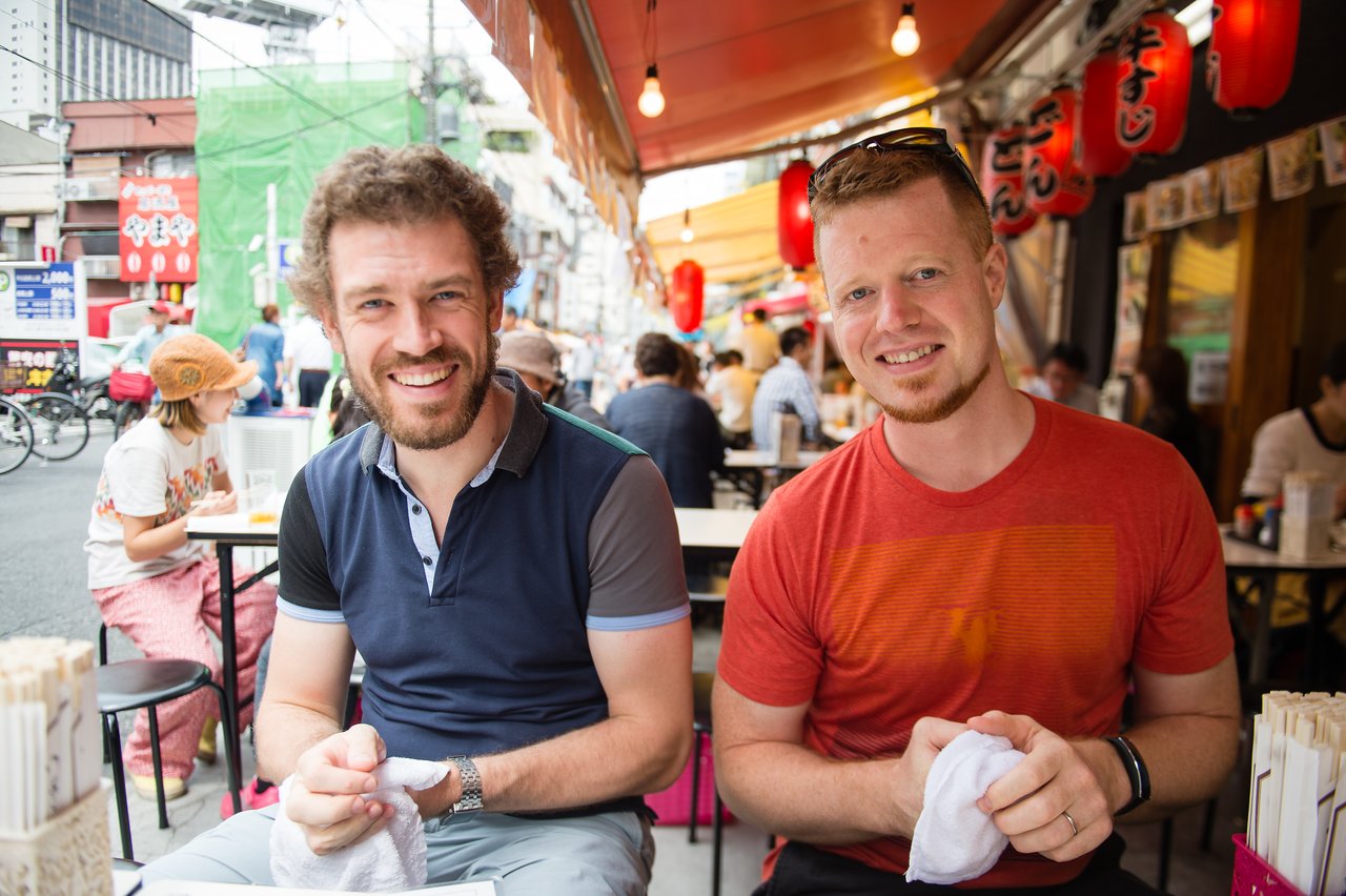 Two men sitting at an outdoor restaurant in Japan, smiling and holding small towels.
