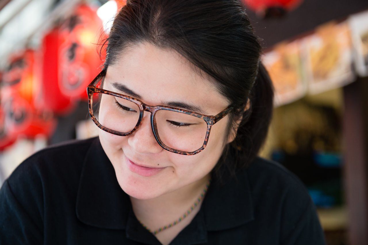 A woman wearing glasses and a black shirt smiles while looking down, with red lanterns in the background.