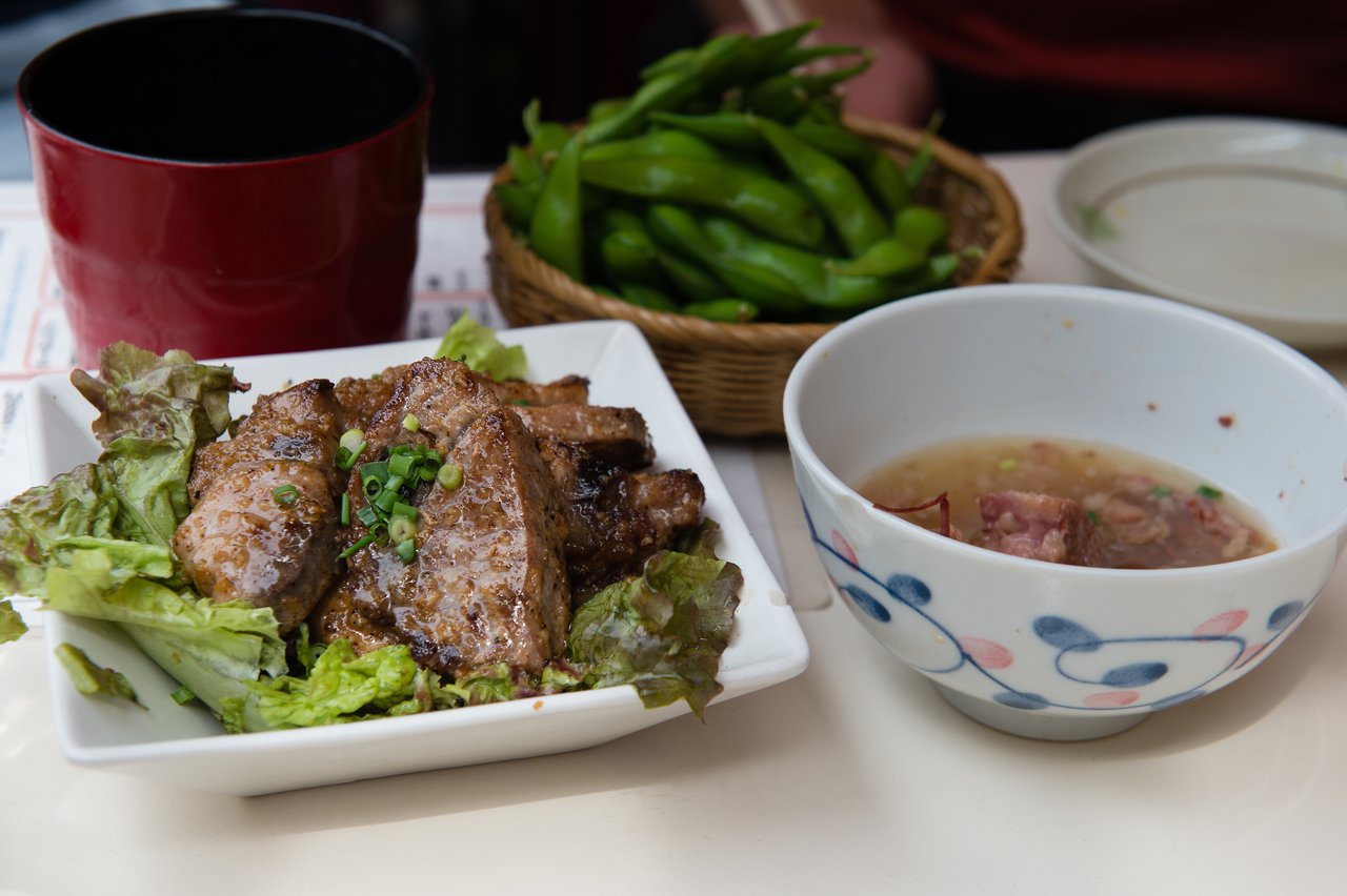 A meal with grilled meat on lettuce, a bowl of soup, edamame, and a red cup on a table.