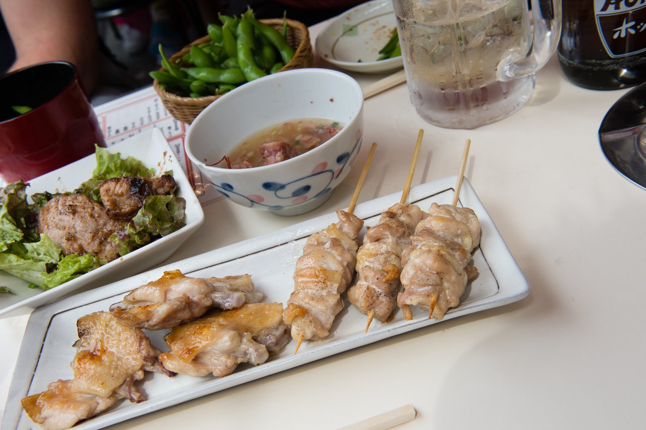 Grilled chicken skewers on a white plate, with salad, soup, edamame, and drinks on a table.