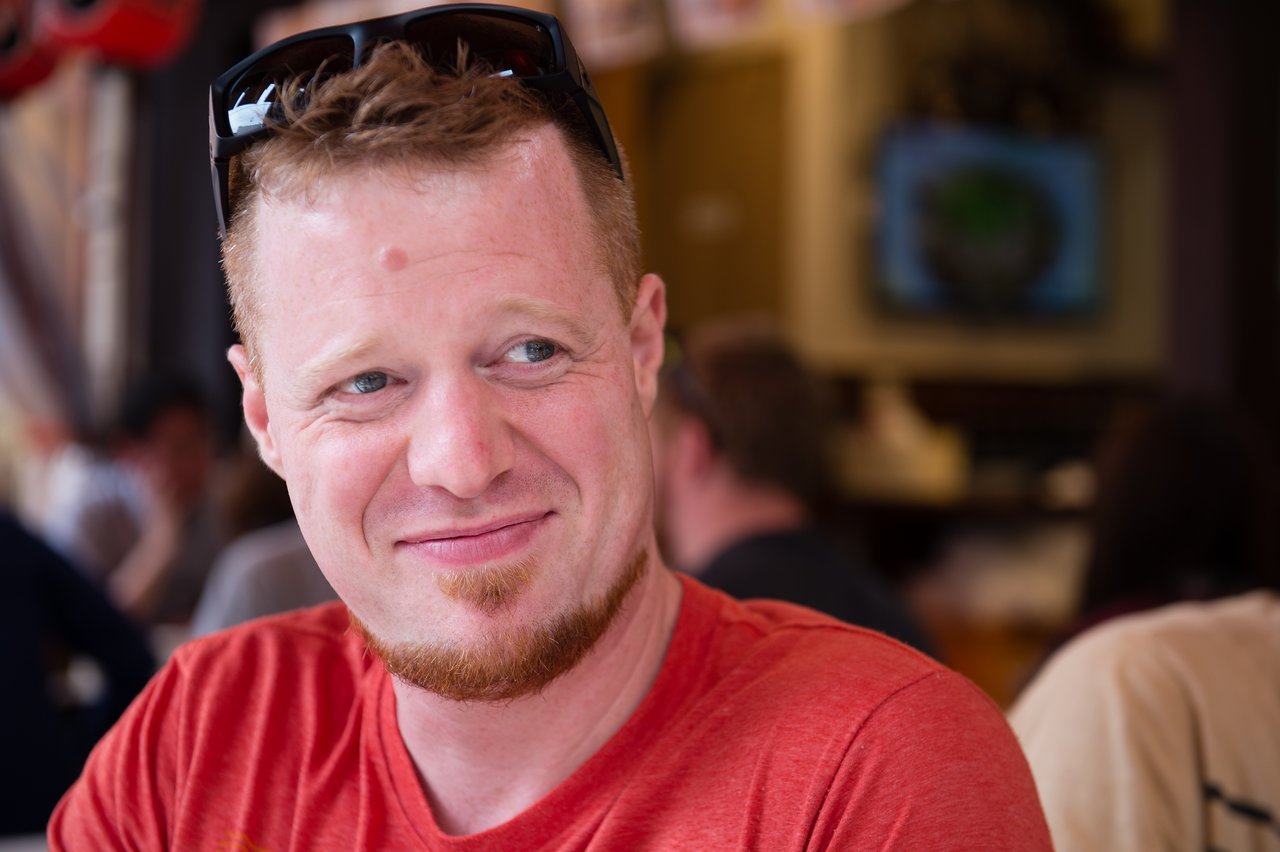 A man in a red shirt smiles while sitting in a busy restaurant.
