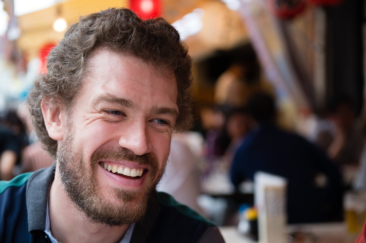 A man with curly hair and a beard is smiling in a busy indoor setting with people in the background.