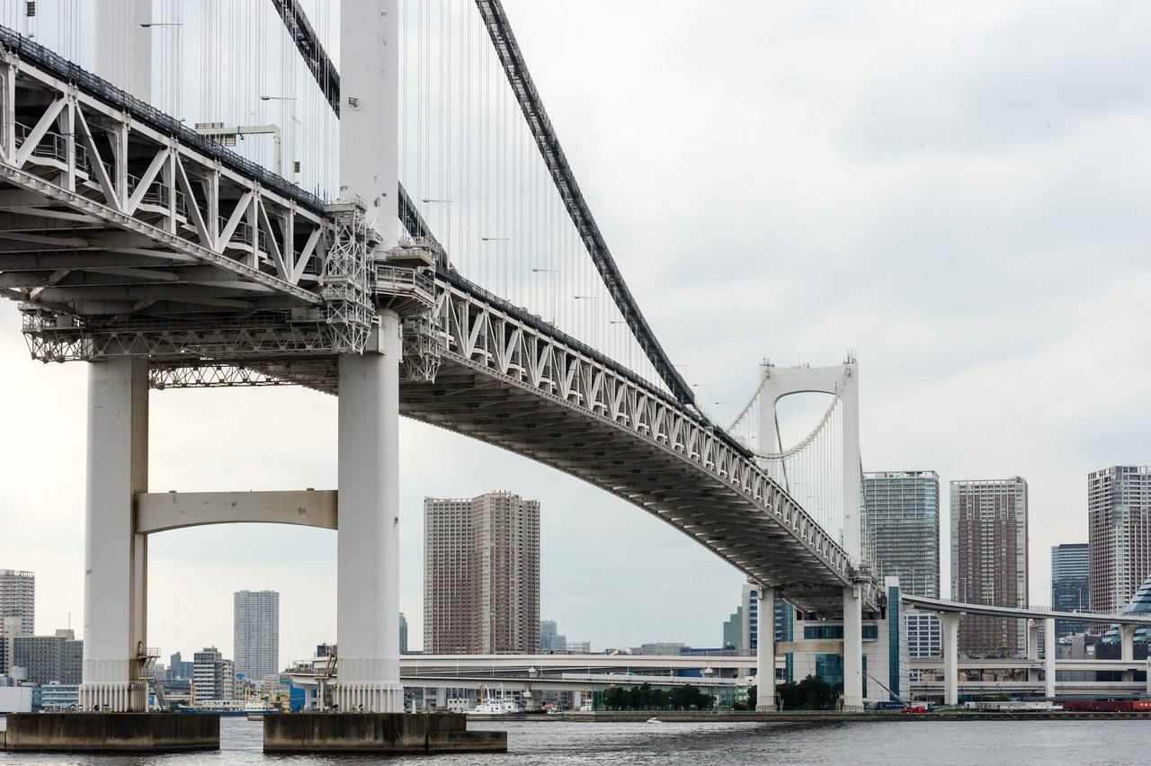 A large suspension bridge spans across the water with tall buildings in the background under a cloudy sky.