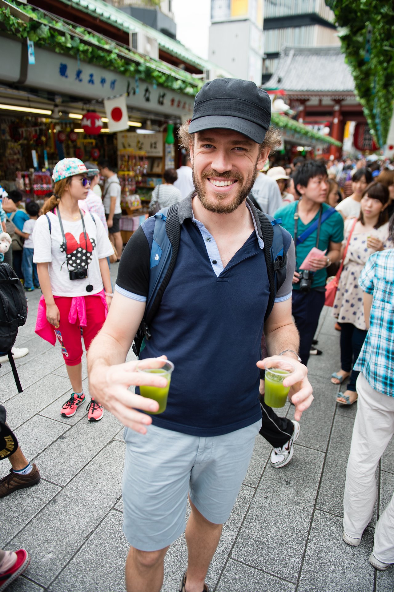 A smiling man in a black cap holds two cups of green tea in a busy Japanese market.