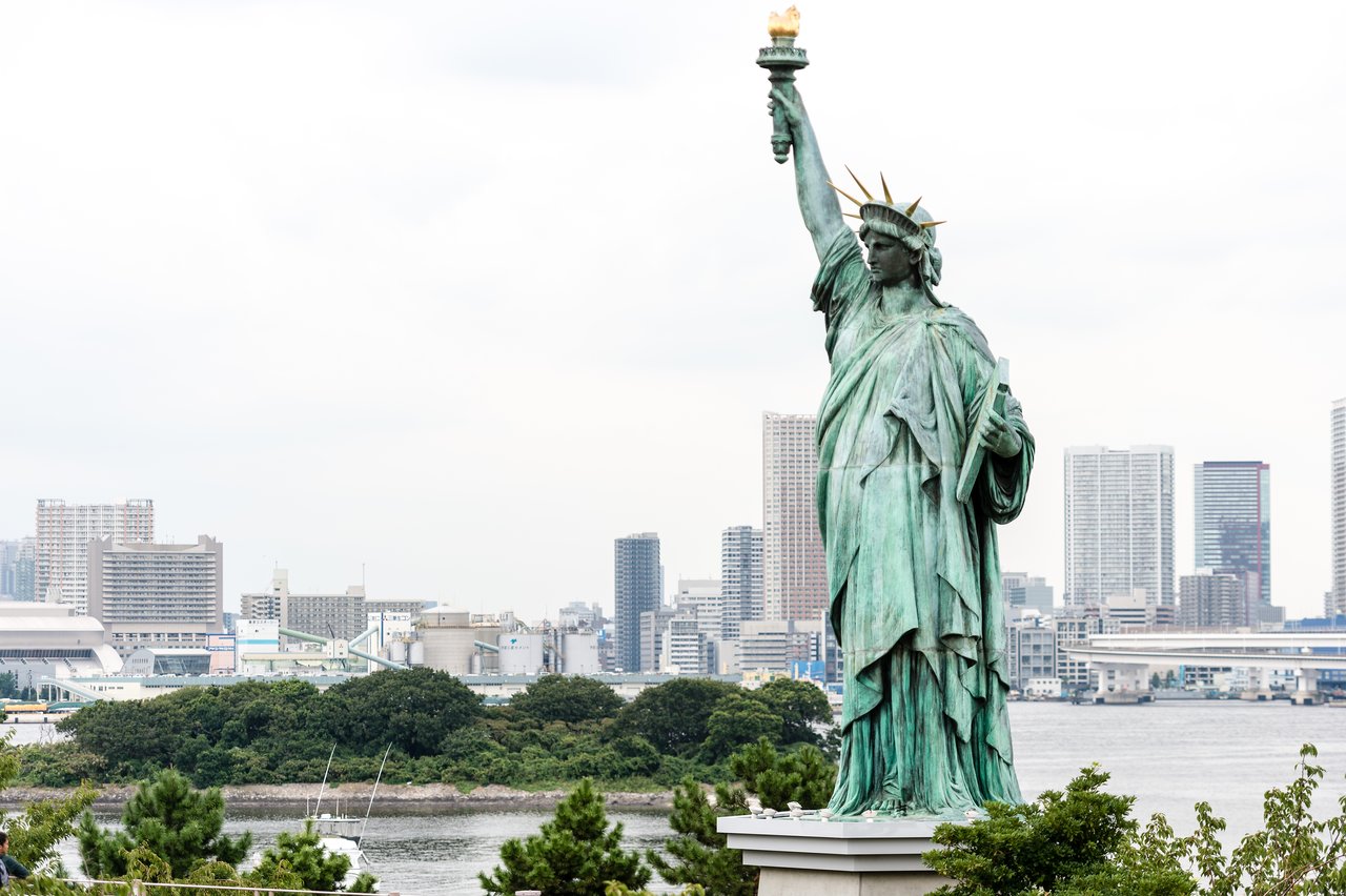 A Statue of Liberty replica stands in front of a city skyline with water and greenery in the foreground.