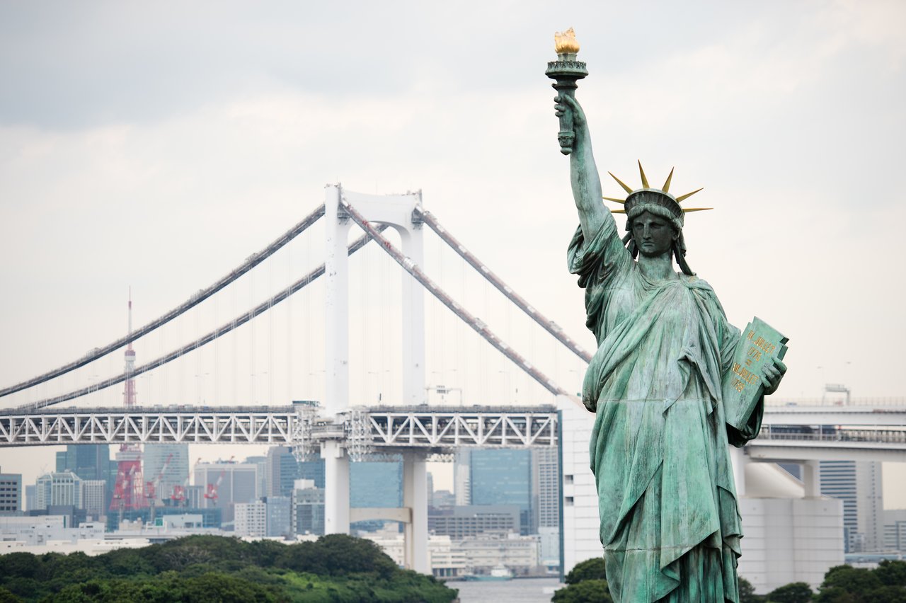 A Statue of Liberty replica stands in front of a large suspension bridge and city buildings in Japan.