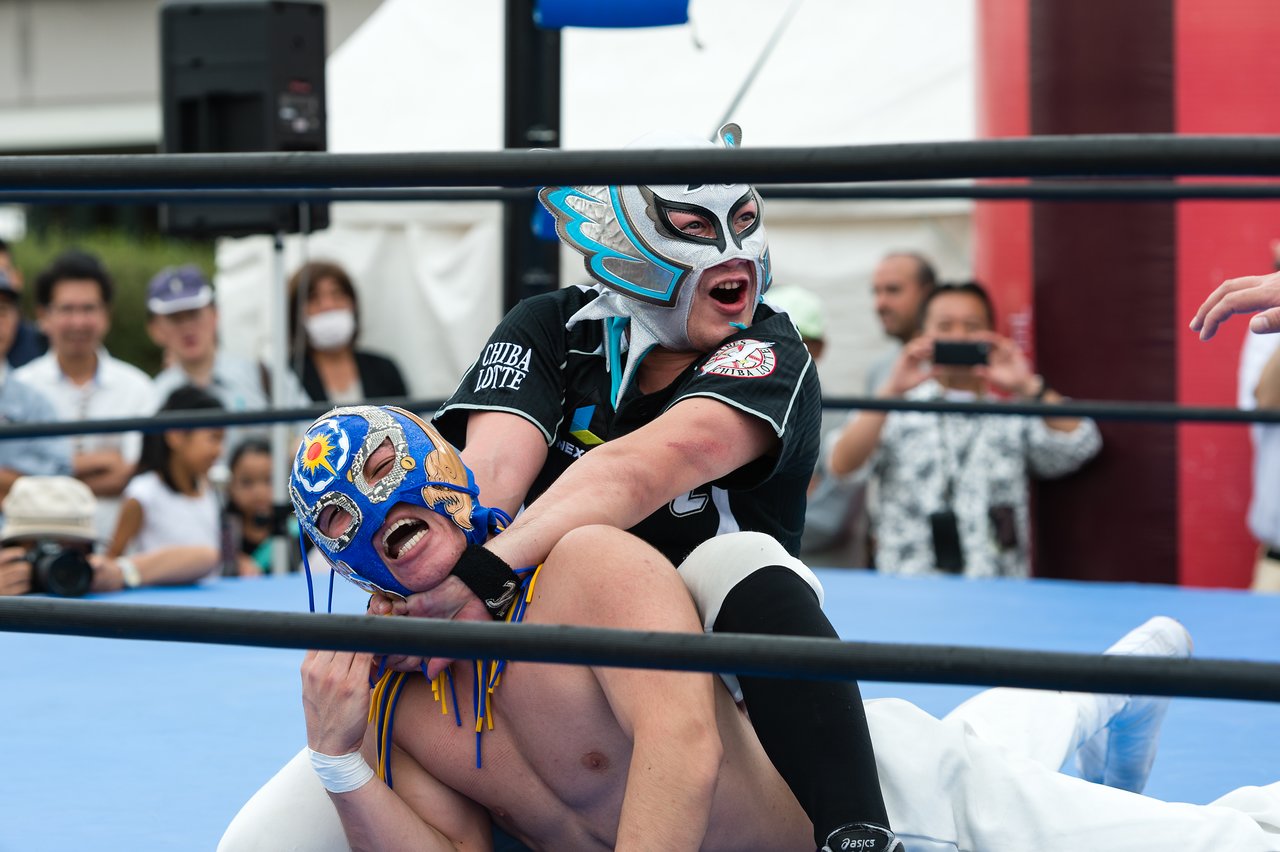 Two masked wrestlers compete in a ring, with one applying a chokehold while a crowd watches.