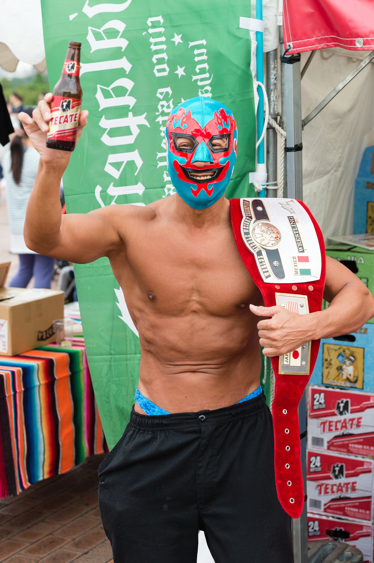 A masked wrestler holds a championship belt and a beer bottle, posing in front of a green banner.