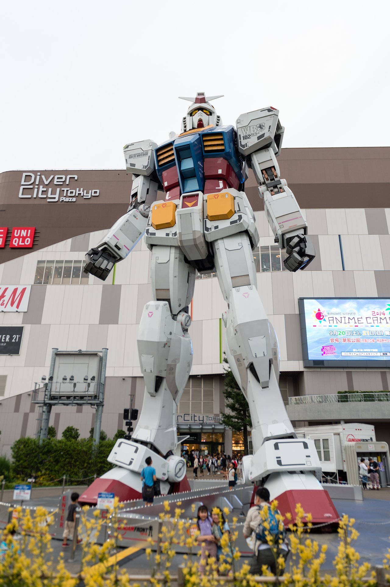 A large Gundam statue stands in front of DiverCity Tokyo Plaza, with people walking nearby.
