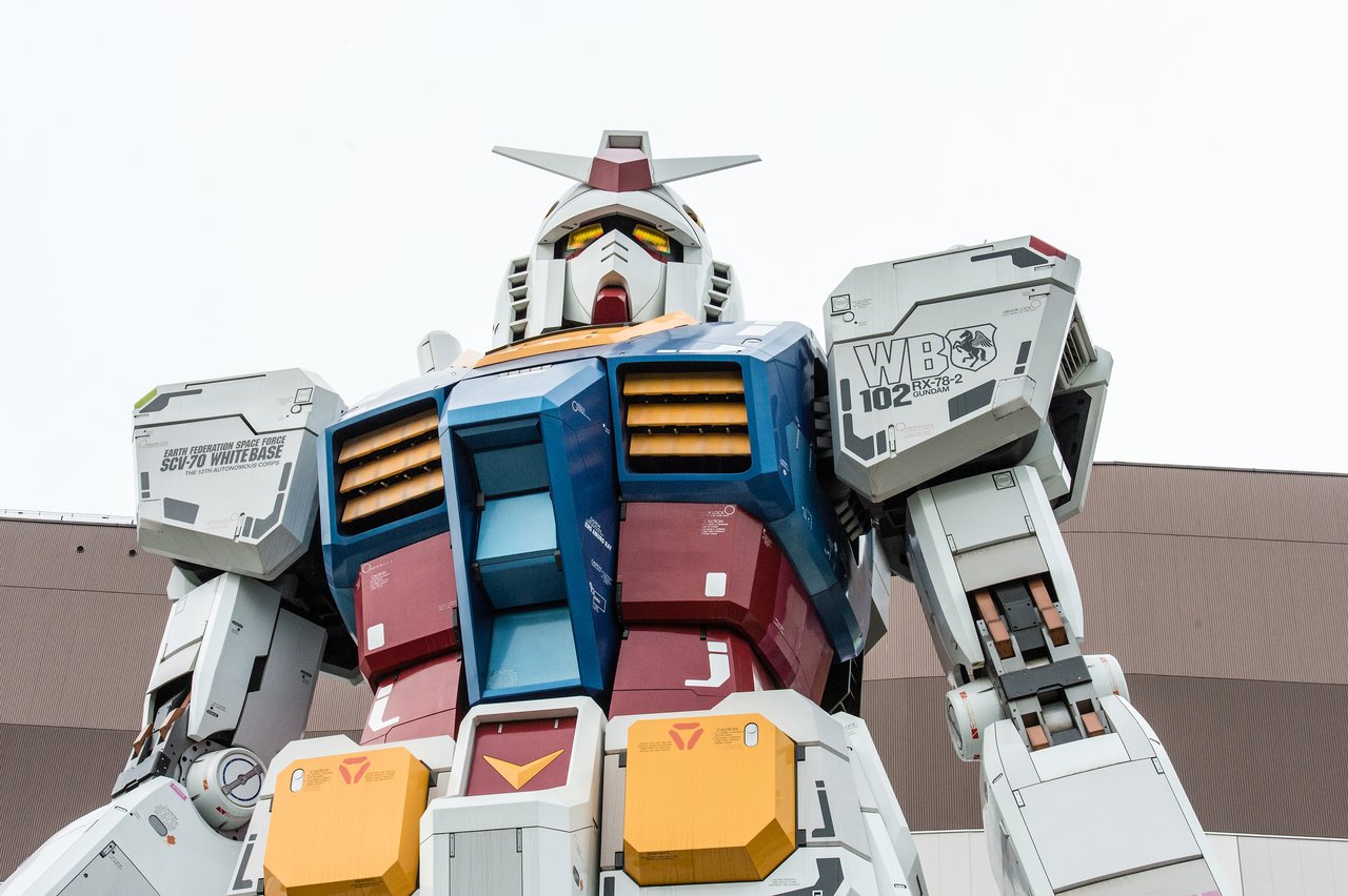 A large Gundam robot statue stands outdoors, featuring detailed mechanical parts and colorful armor.