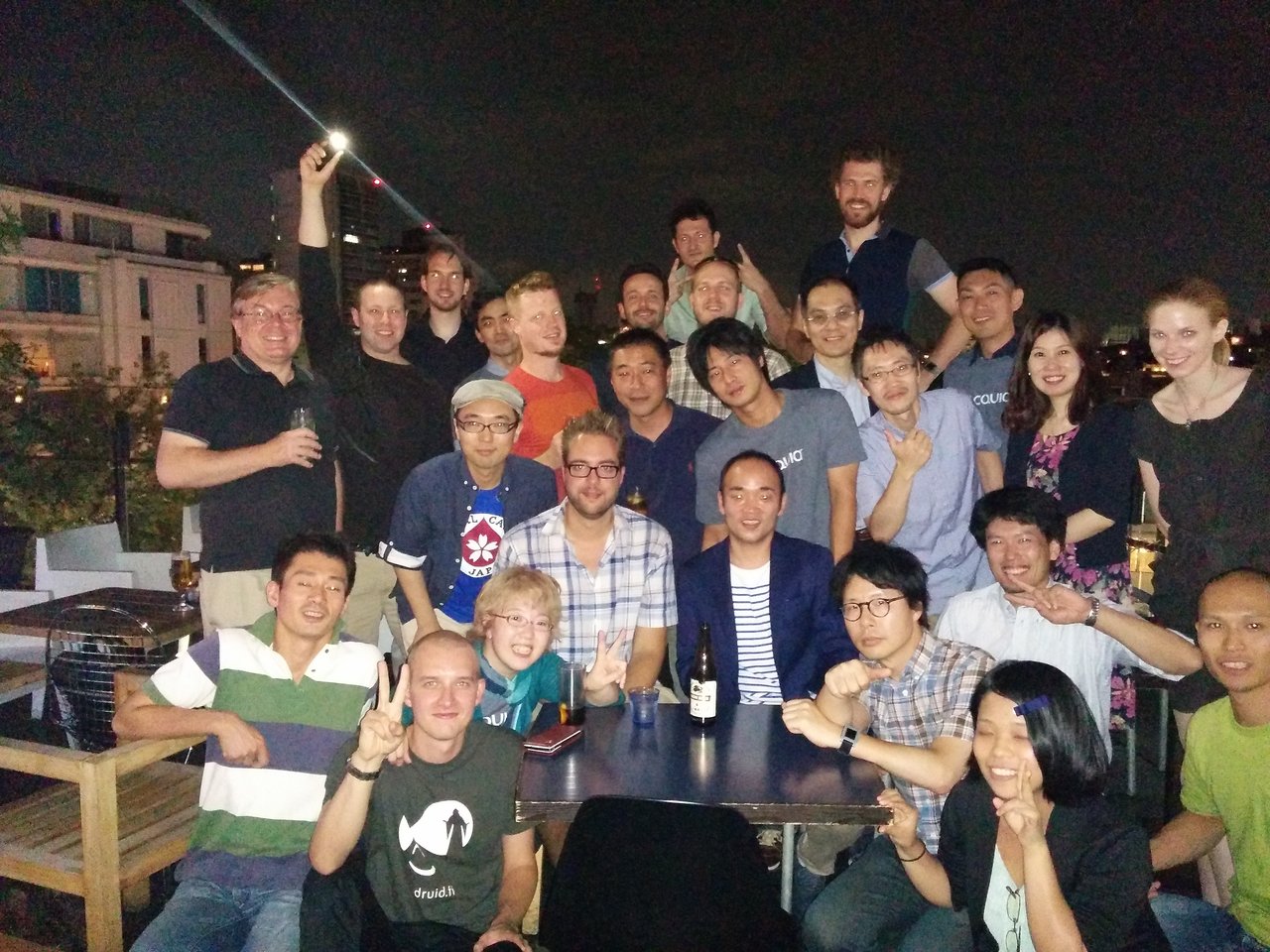 A large group of people posing together at an outdoor gathering, smiling and making hand gestures.