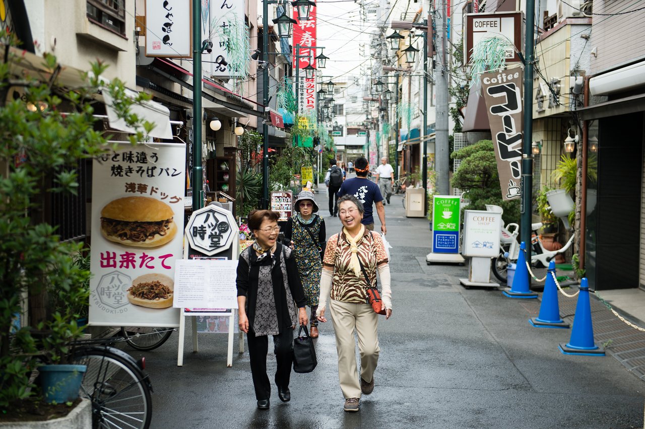 Two women walk and smile on a narrow street lined with shops, signs, and restaurants in Japan.
