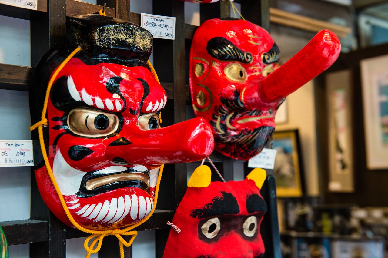 Two red Tengu masks with long noses hang on display in a shop, alongside a smaller red fabric mask.