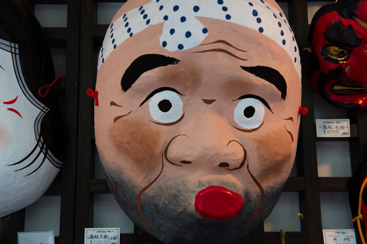Large traditional Japanese mask with a surprised expression, red lips, and a polka-dotted headband, displayed on a wall.