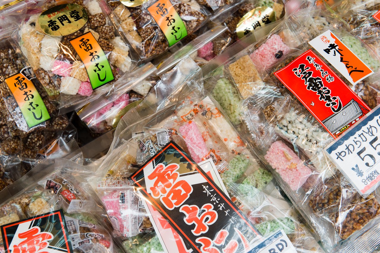 Bags of colorful Japanese rice crackers in clear packaging with bold labels displayed for sale at a market.