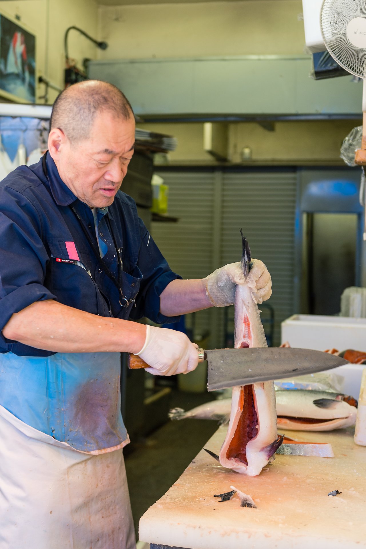A fishmonger wearing gloves and an apron cuts a large fish in half with a cleaver.