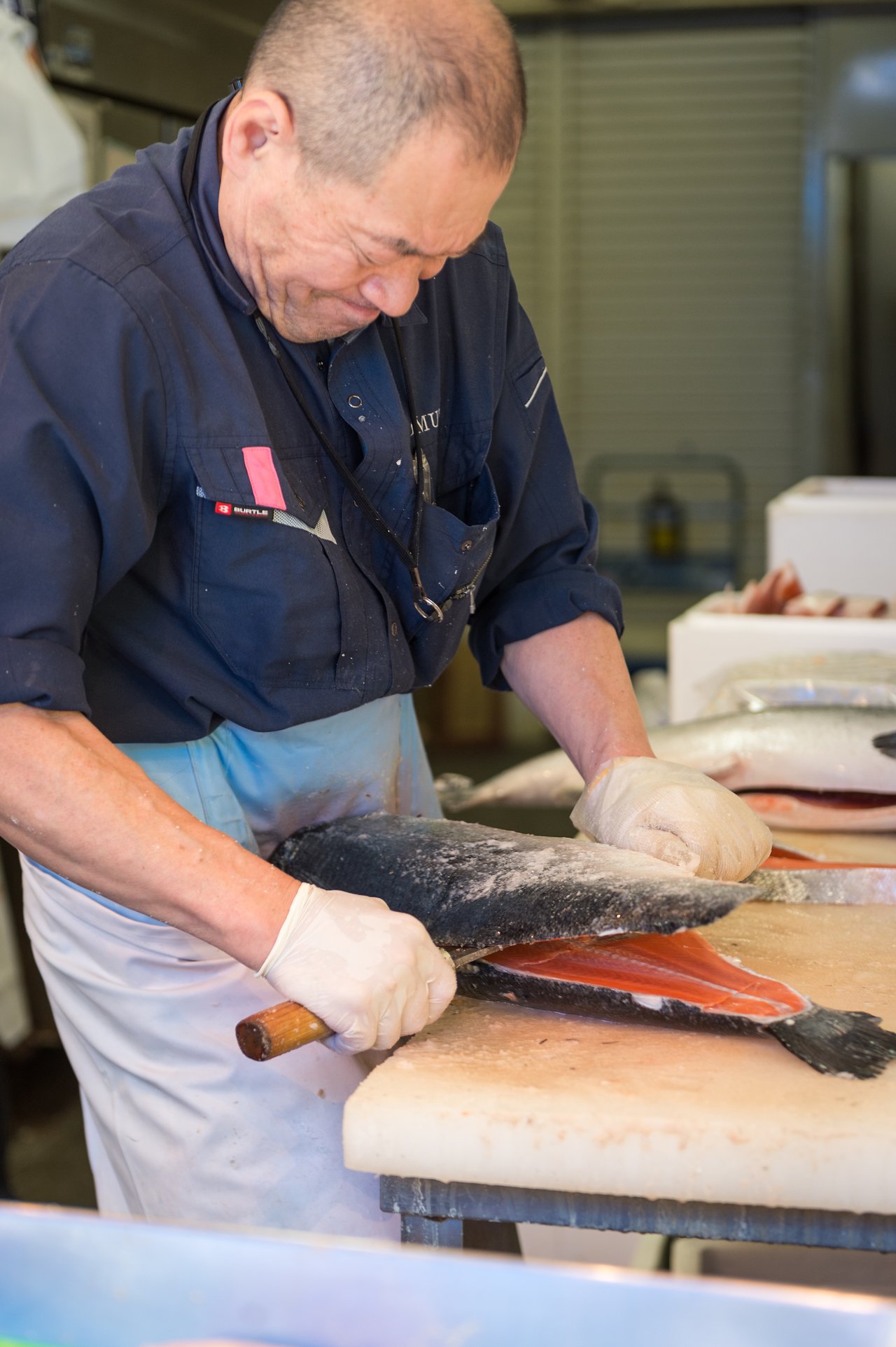 A fishmonger wearing gloves fillets a large salmon on a cutting board using a sharp knife.