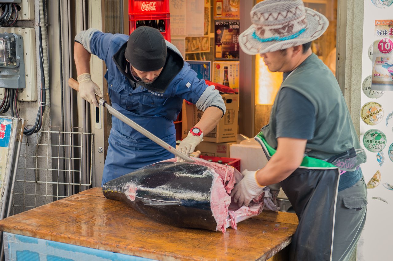 Two workers cut a large tuna fish on a wooden table using a long knife.