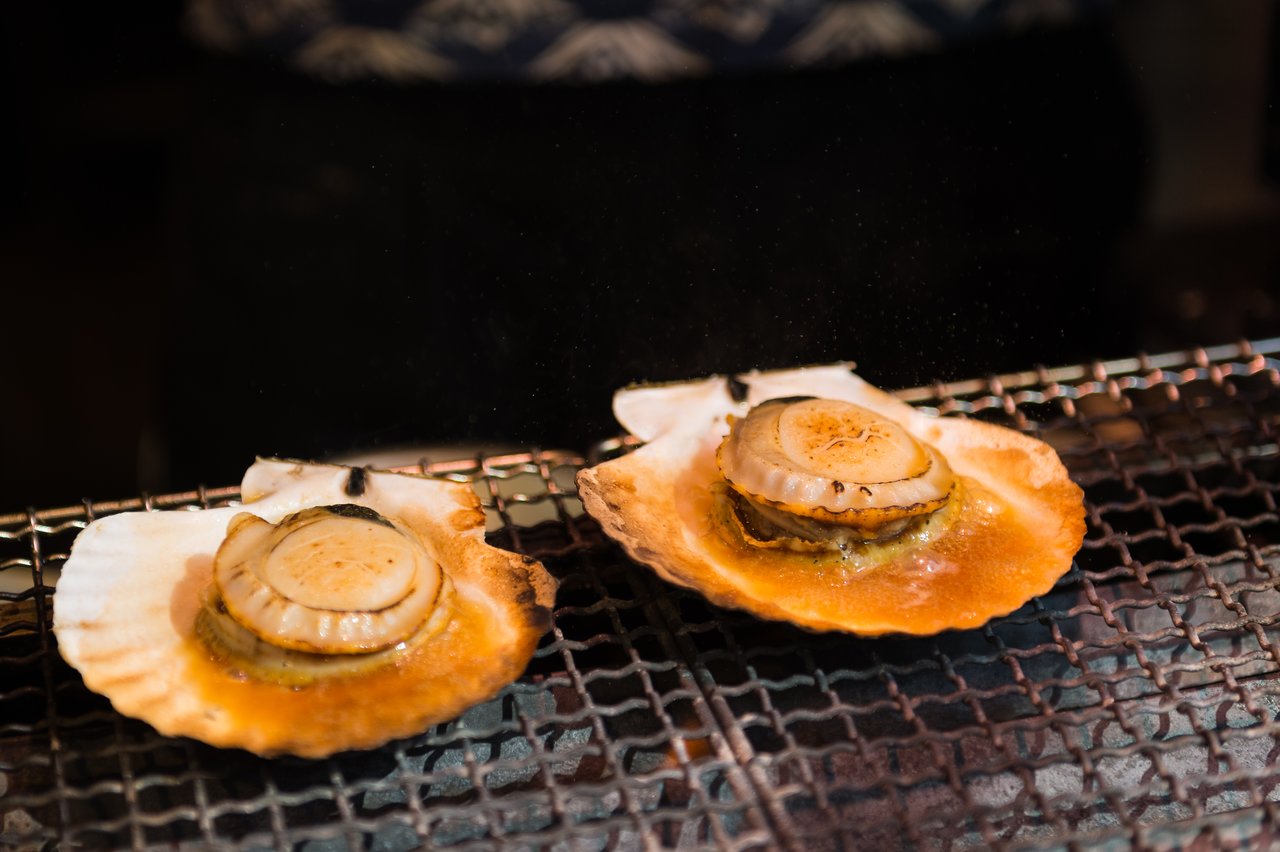 Two scallops in their shells are grilling over an open flame, with juices sizzling on the hot surface.